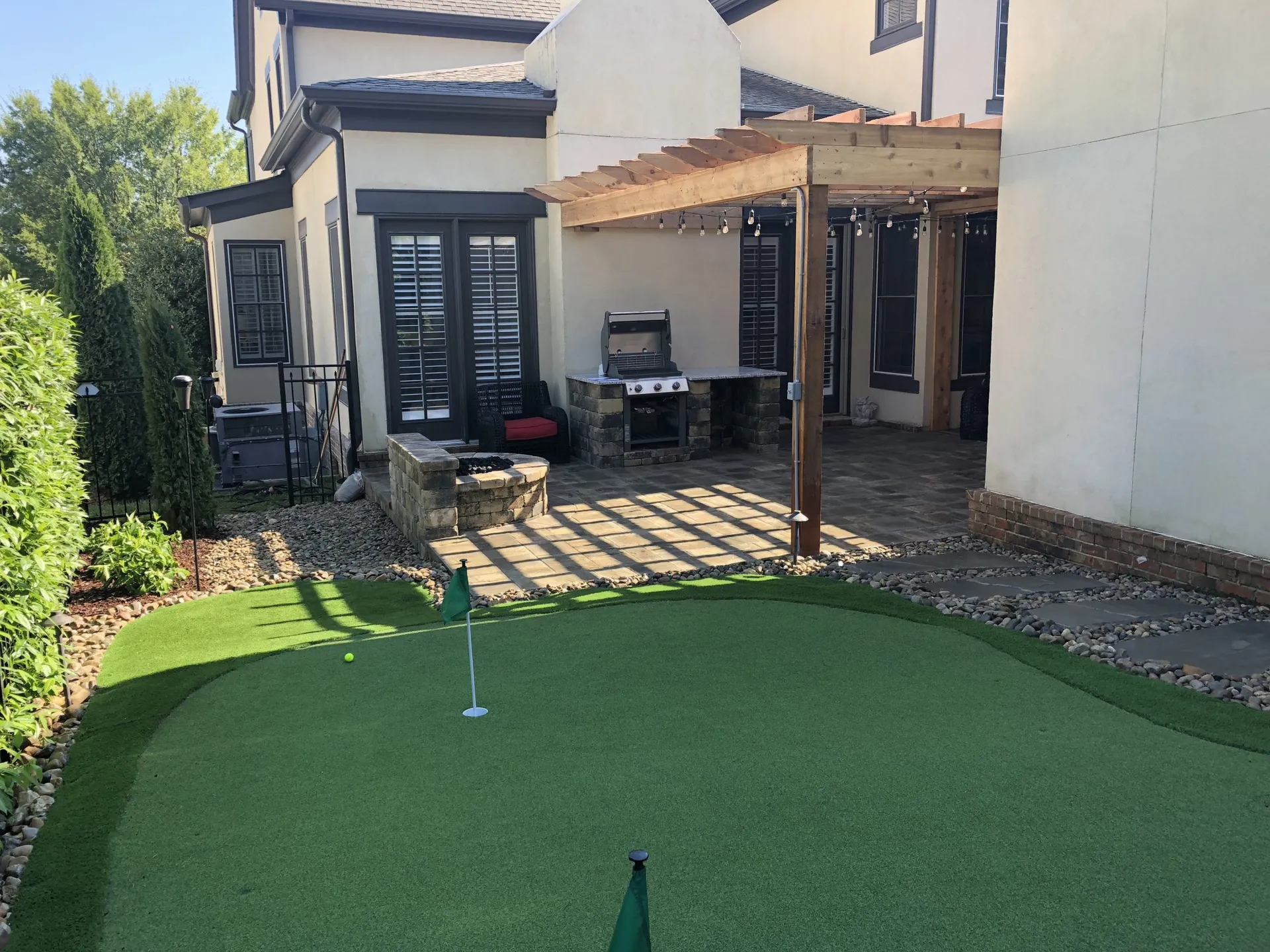 Artificial turf shaped into a putting green with a flag in a backyard, next to a stone fire pit, a grill and a wood pergola beside a cream stucco house.