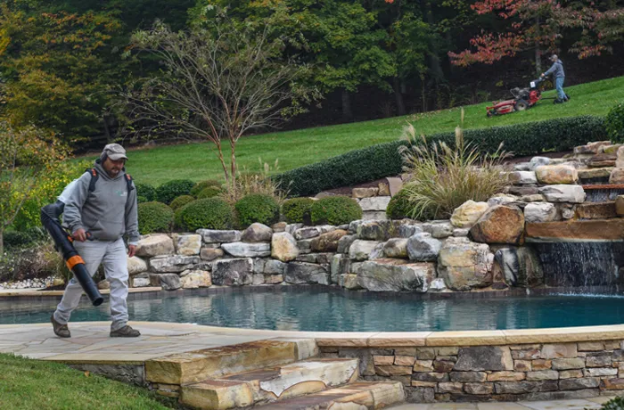 A Green Scene crewman with a backpack blower clearing clippings off the stone edge of a backyard pool while another mows the slope above
