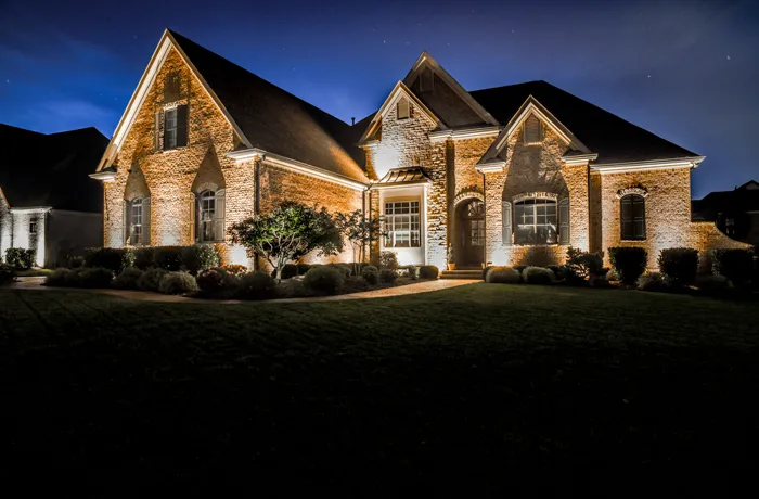 A two-story brick home in Brentwood at dusk, its gables and arched windows washed in warm uplighting above dark shrub beds and a front lawn