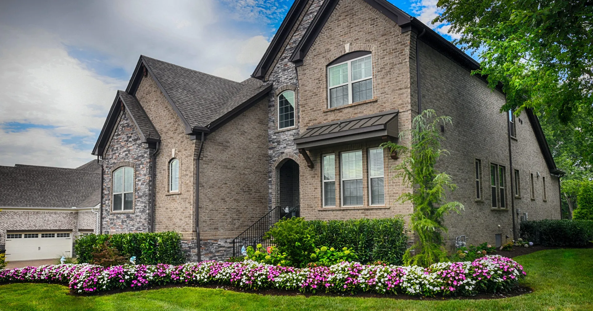 A two-story brick and stone home with a curving bed of pink and white flowers across the front lawn and shrubs along the foundation