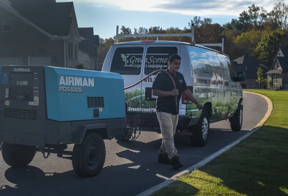 A Green Scene technician walking beside a towed air compressor hitched to the company van on a curving neighborhood street