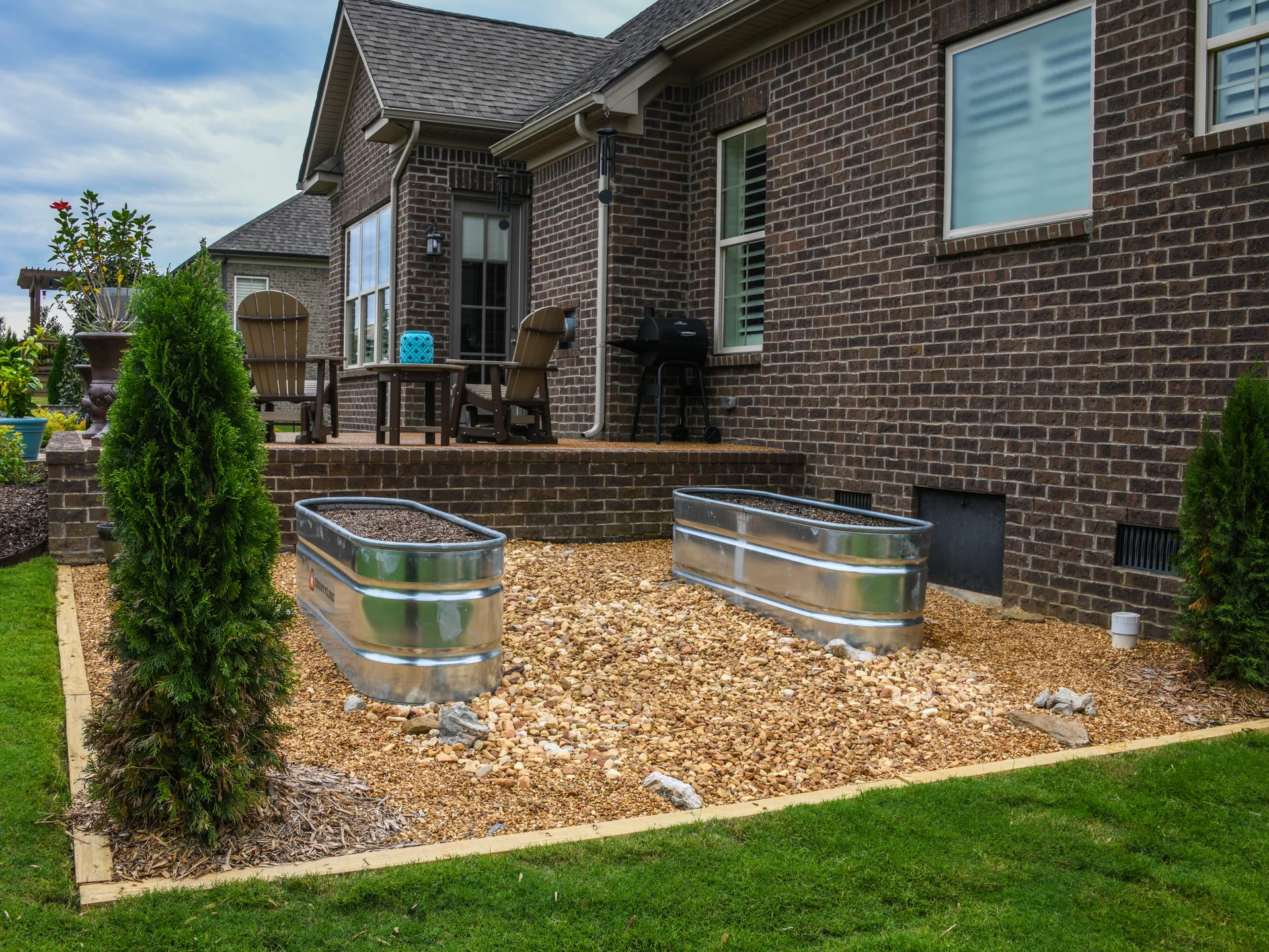 Galvanized container garden beds in front of a row of young arborvitae in a backyard