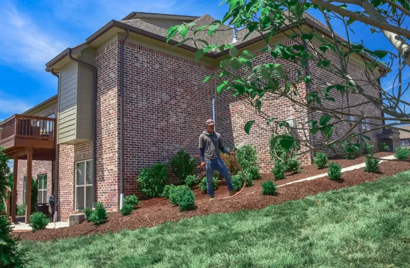 A Green Scene crewman watering newly planted shrubs in a fresh mulch bed on the slope beside a brick home.