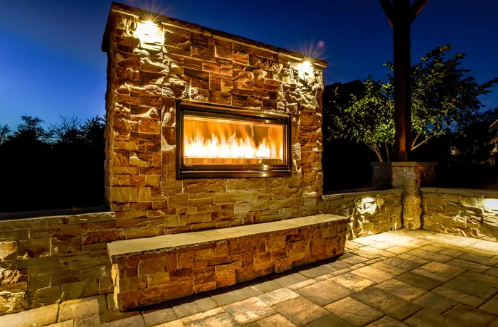 A paver patio around a stone outdoor fireplace with a gas insert, lit at night