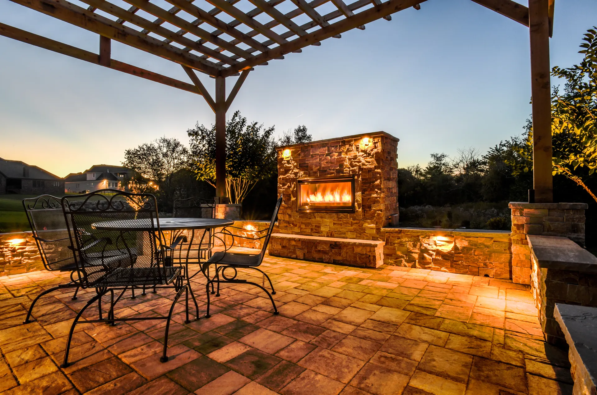 A stone gas fireplace lit at night under a wood pergola, with an iron bistro table and chairs on a paver patio lit by seat-wall lights.