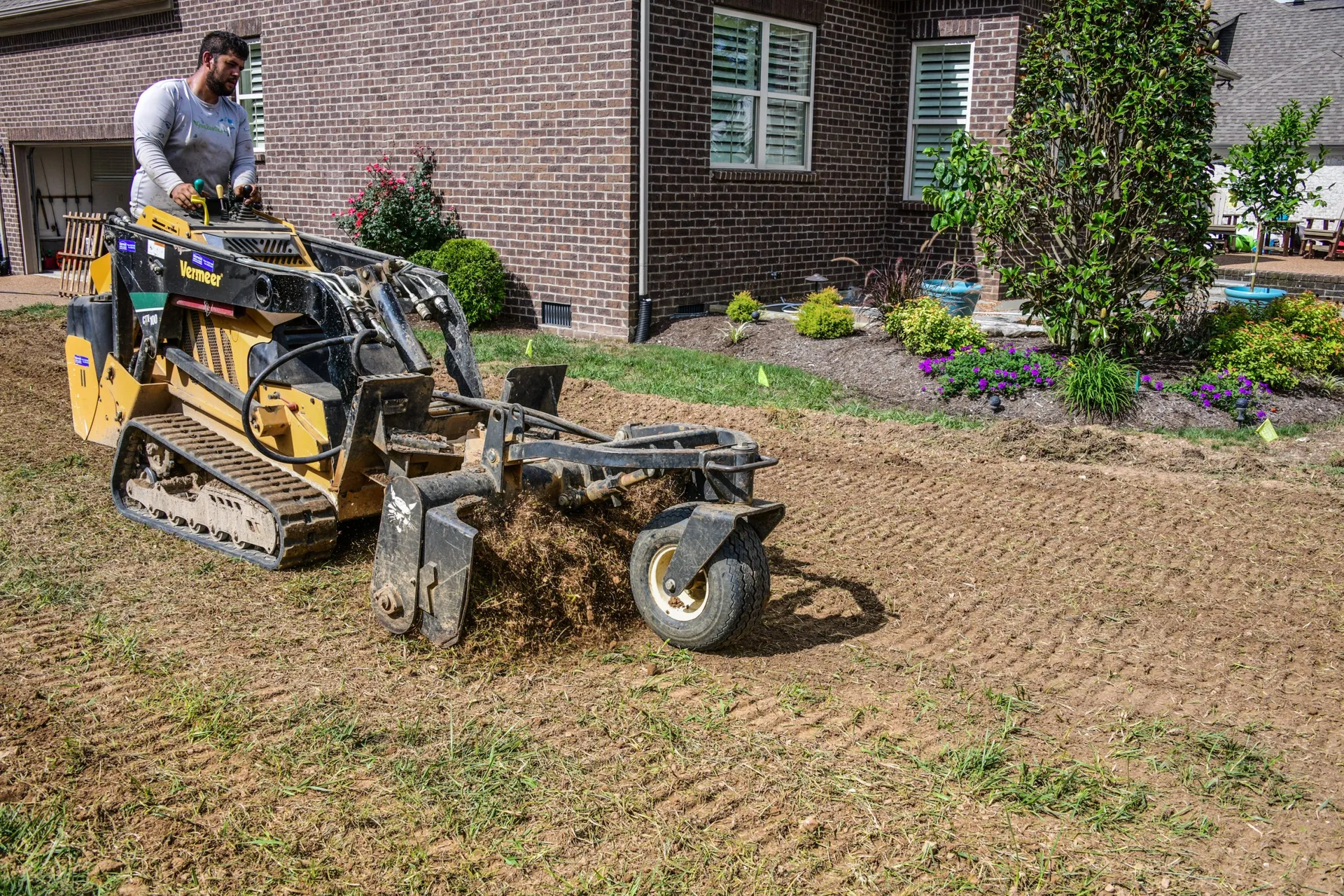 A Green Scene operator on a stand-on skid steer running a Harley rake across bare soil beside a brick house