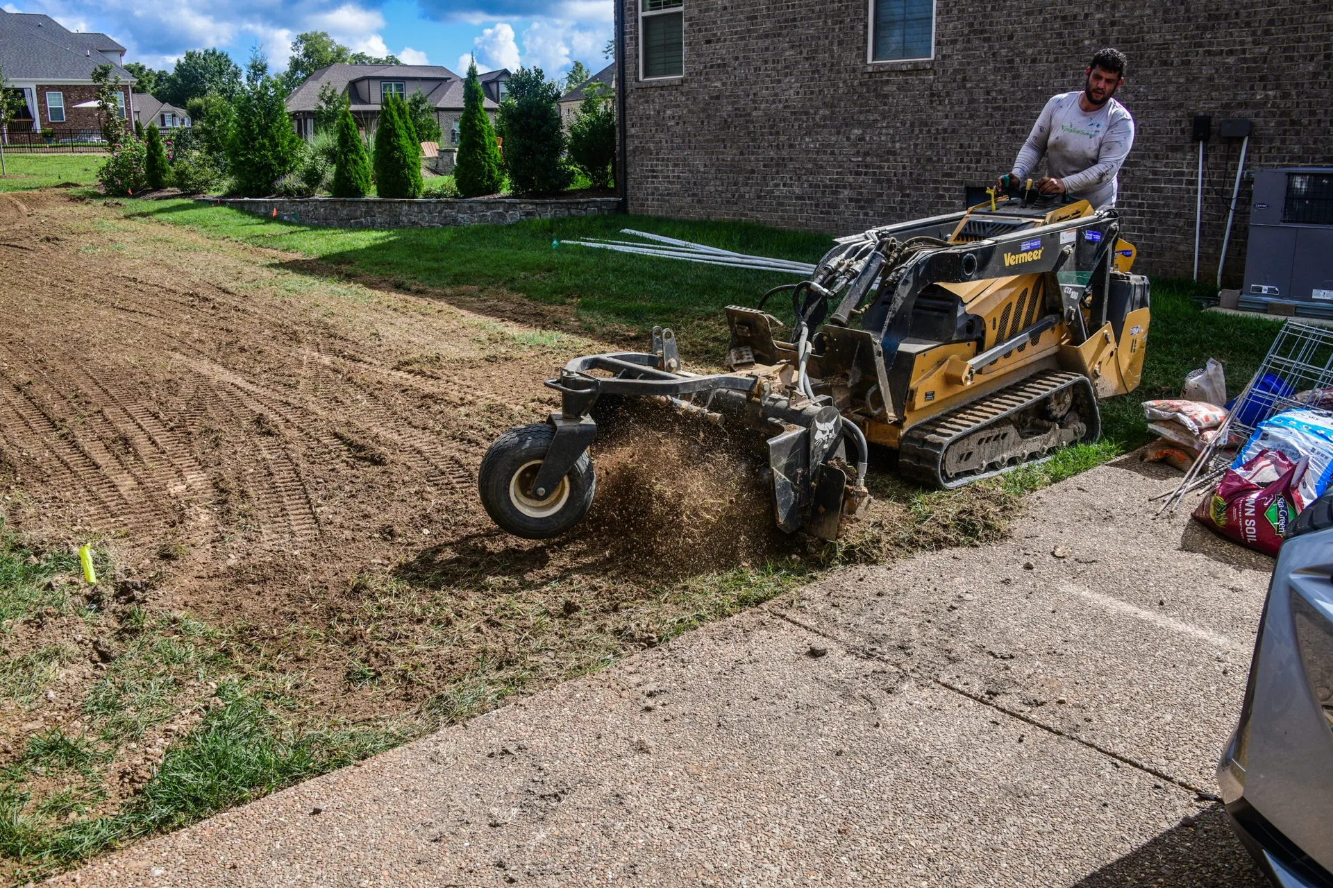 A Harley rake on a stand-on skid steer churning up old turf at the edge of a driveway, throwing a spray of soil
