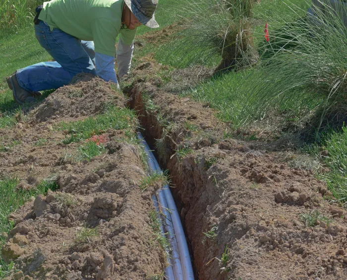 A crew member kneeling beside an open trench with new irrigation pipe lying in the bottom