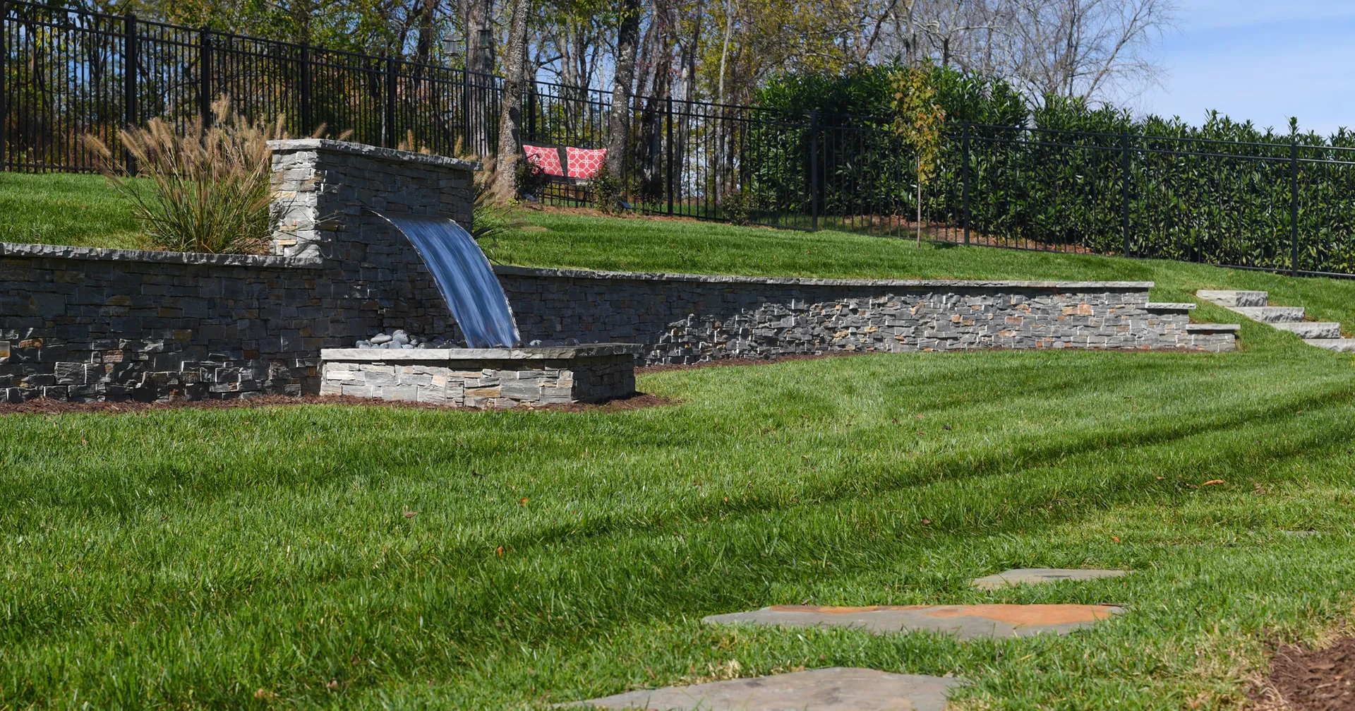 A terraced backyard lawn held by stacked-stone retaining walls, with a sheer stream of water pouring from the upper wall into a stone basin and stone steps climbing the slope.