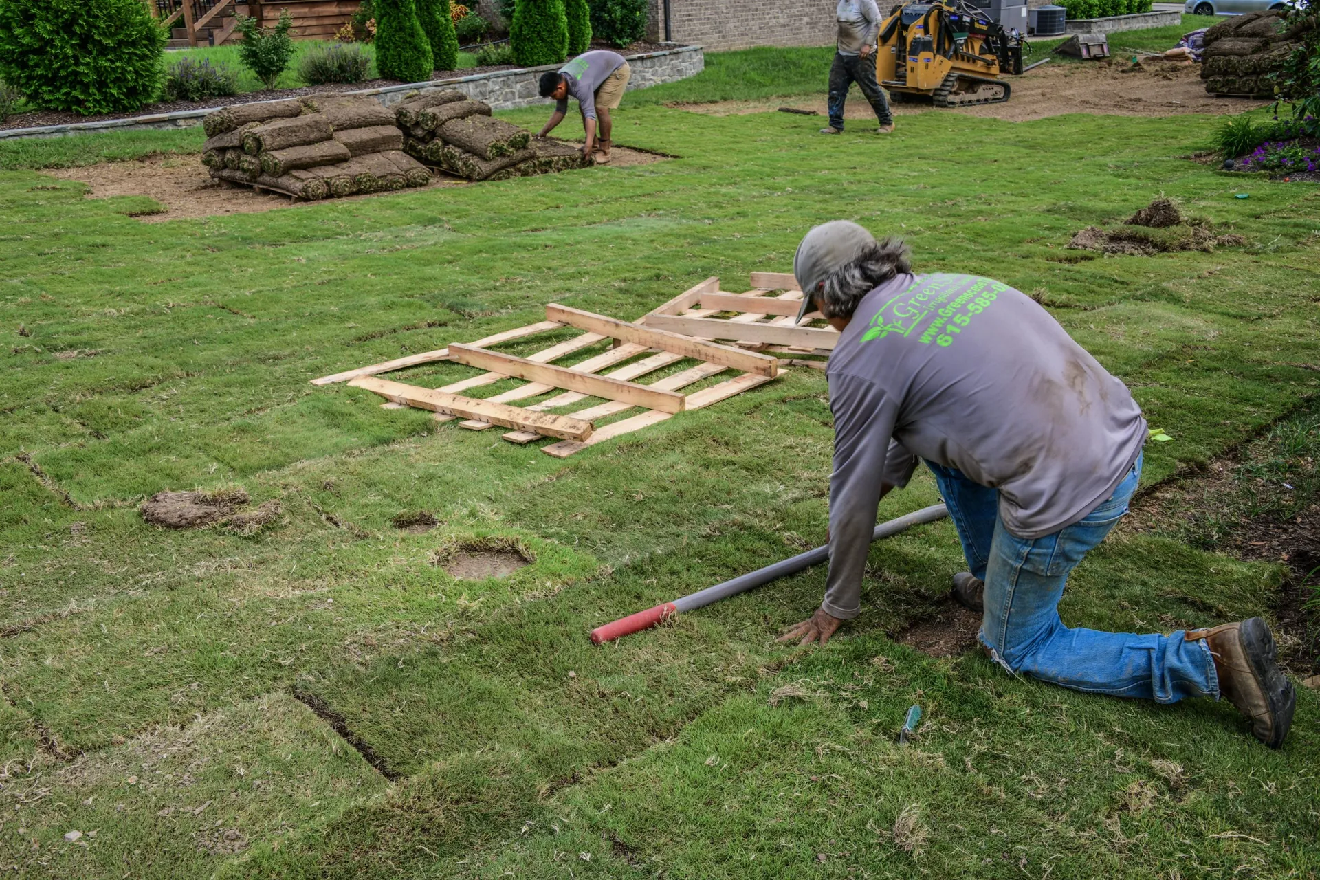 A Green Scene crewman on his knees fitting a roll of fresh sod into place, with stacked rolls and a compact loader behind him