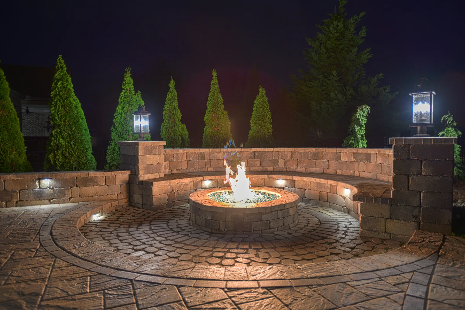 A round fire pit burning at night inside a curved stone seat wall, with small lights set into the wall, lanterns on two stone pillars and a circle of cobble pavers around the pit.