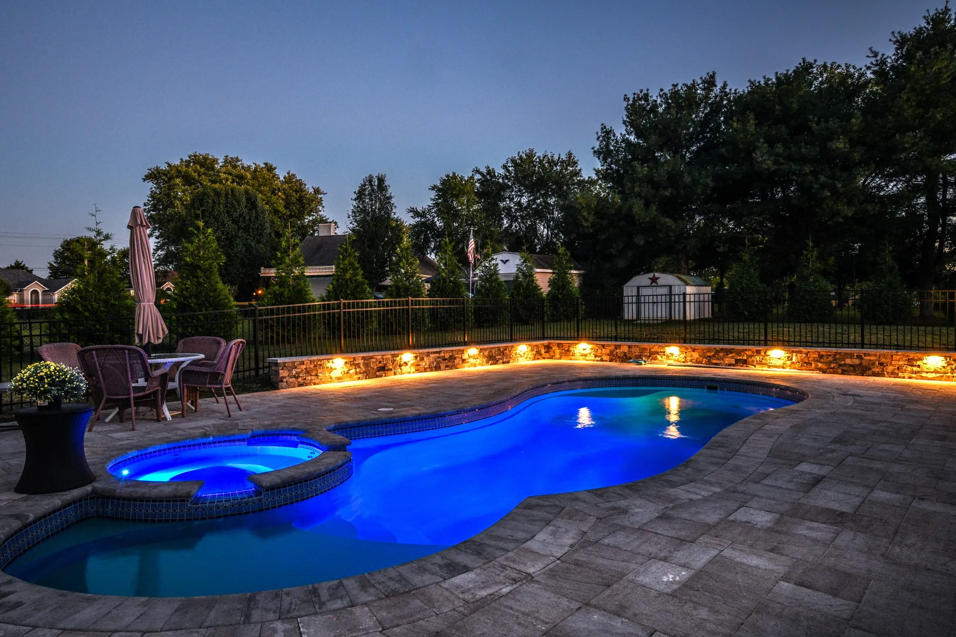 A pool lit blue at dusk, ringed by a gray paver deck, with a stone wall behind it lit along its face and a table and umbrella at the far end.