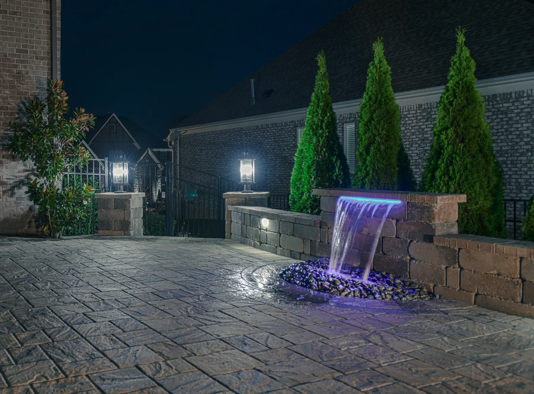 A sheet of water lit blue at night, falling from a stone seat-wall pillar onto black river rock beside a patio and a row of tall evergreens.
