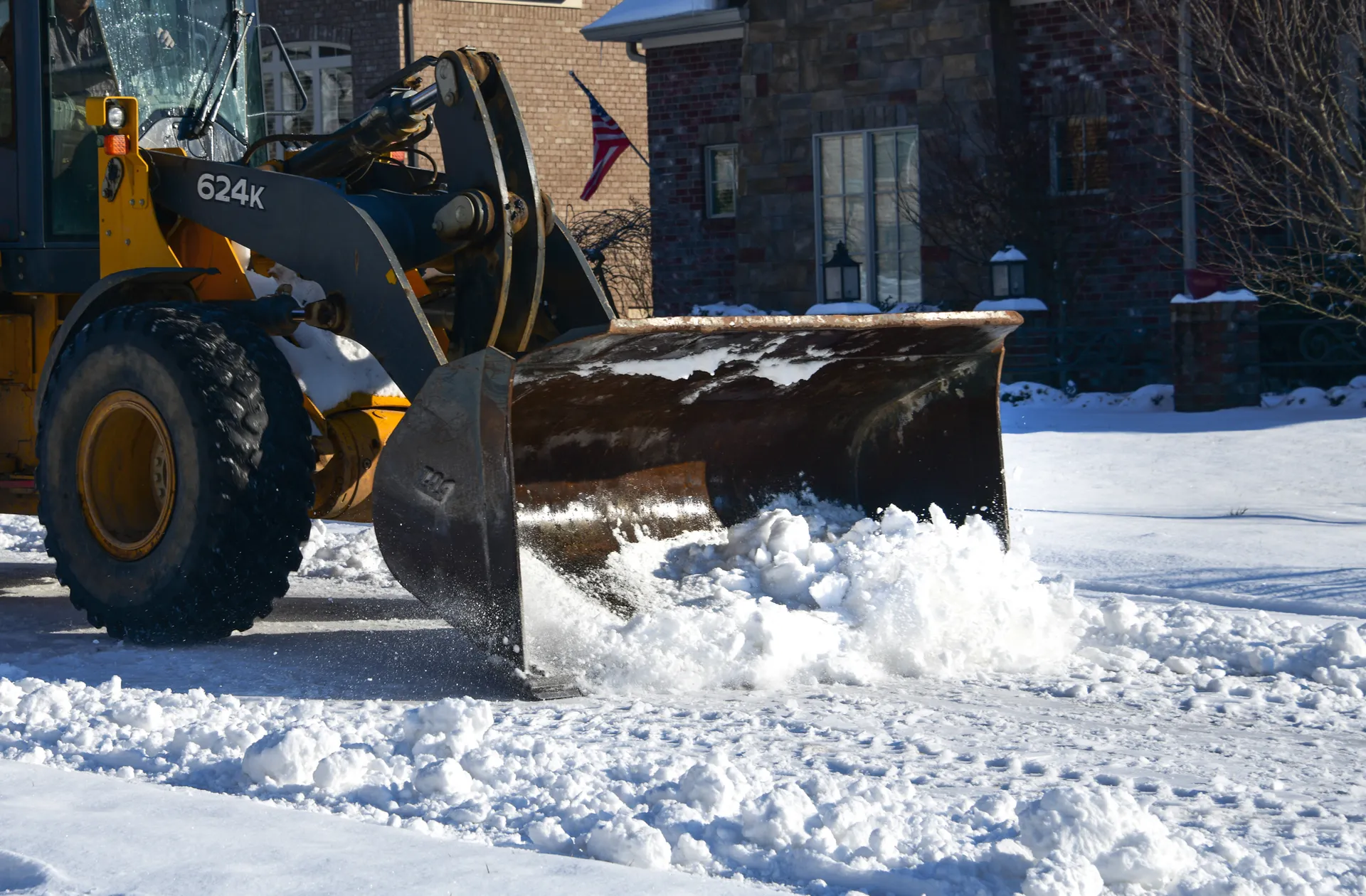 A yellow wheel loader with a plow blade pushing snow off a driveway in front of a brick house