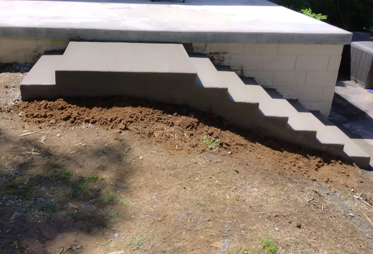 New poured concrete steps rising along a concrete block wall to a porch slab, with fresh soil backfilled along their base.