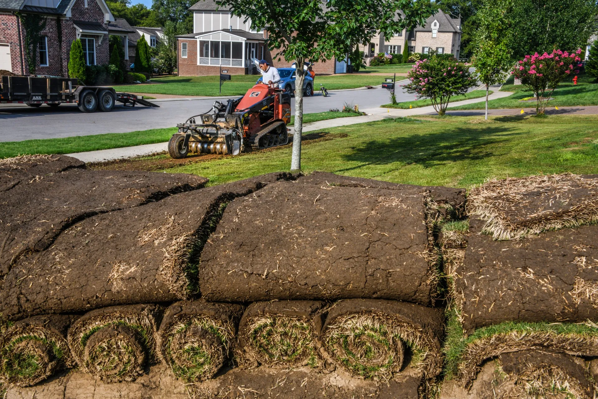 Rolled sod stacked on a pallet at the edge of a lawn, with a compact loader parked by the street