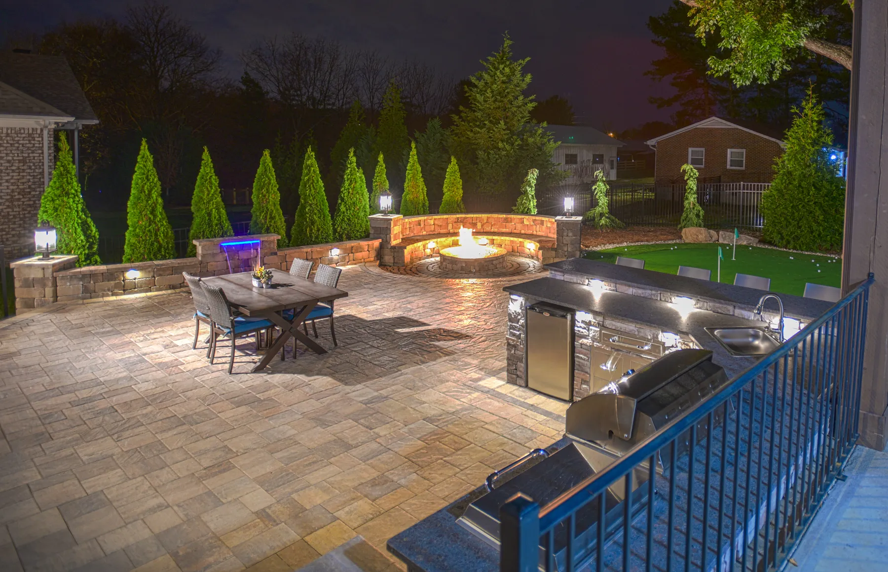 A paver patio at night with a fire pit burning inside a curved stone seat wall, a dining table, an outdoor kitchen with a grill and sink, and a putting green beyond, lit by lights set into the walls and lanterns on stone pillars.