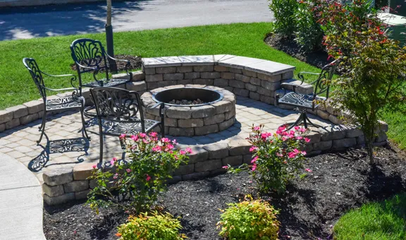 A round stone fire pit on a circular paver patio, partly wrapped by a low curved seat wall, with iron chairs and flowering shrubs in a mulch bed.