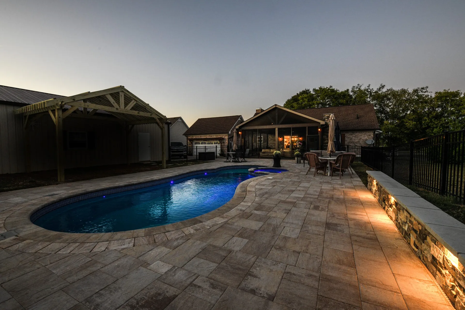A pool lit blue at dusk with a wide paver deck, a lit stone seat wall along the fence, and a pavilion and covered porch beyond.
