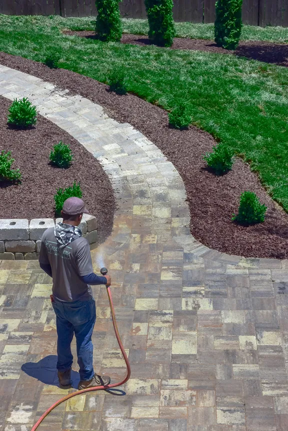 A crewman rinsing a newly laid curved paver walkway that runs between fresh mulch beds and young shrubs.