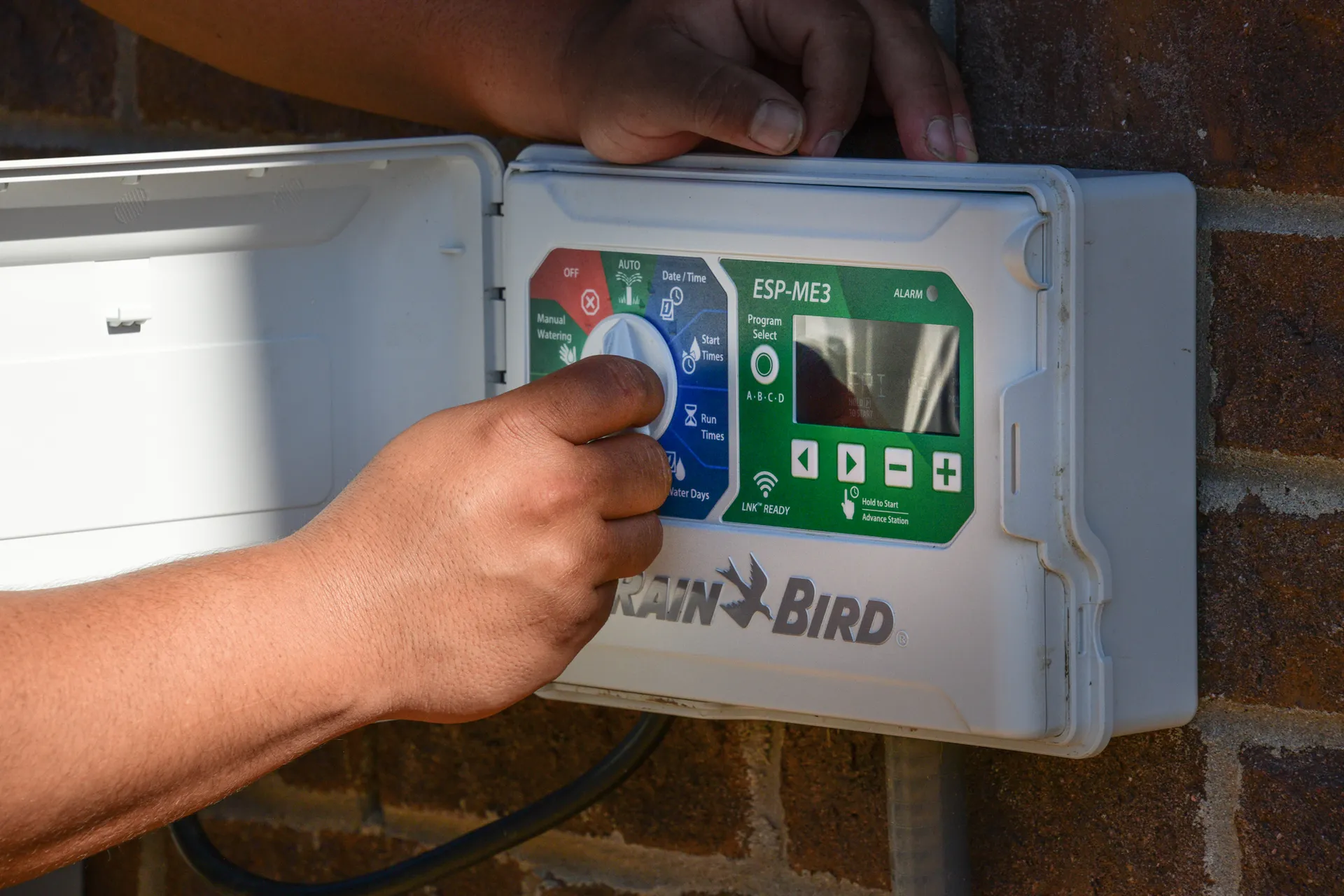 A hand turning the dial on a Rain Bird irrigation controller mounted on a brick wall