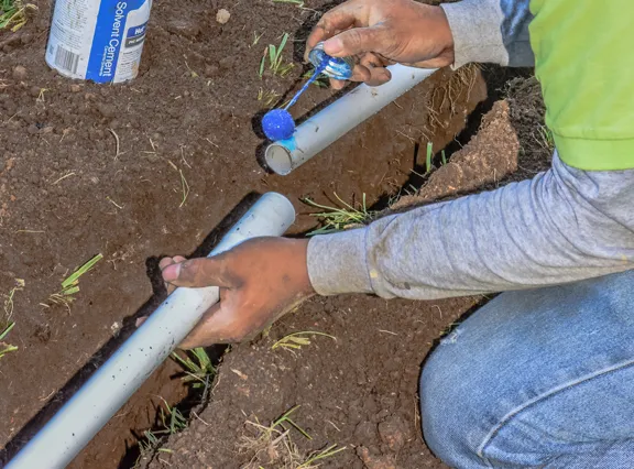 A technician applying solvent cement to a length of white PVC pipe beside a cut line in an open trench