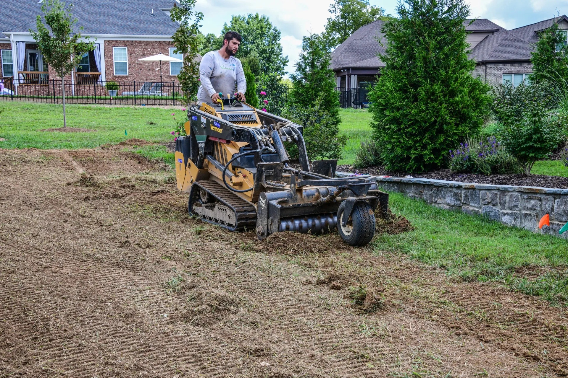 A crewman on a skid steer with a Harley rake grading a raked backyard smooth beside a stone wall and a row of young evergreens