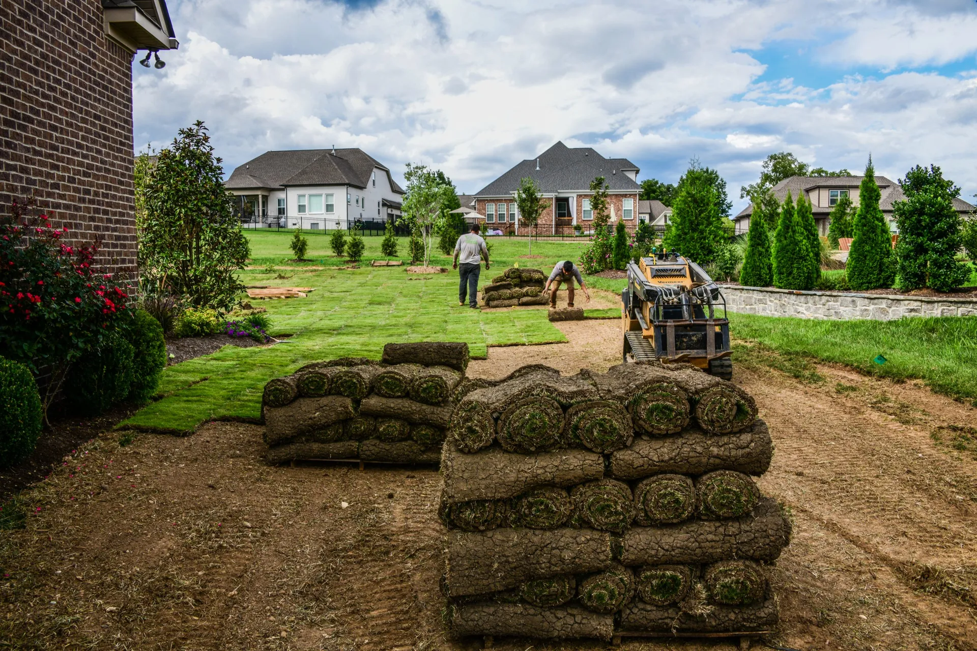 Stacks of sod rolls staged on bare, leveled soil in a backyard, with a skid steer and a finished strip of lawn behind them