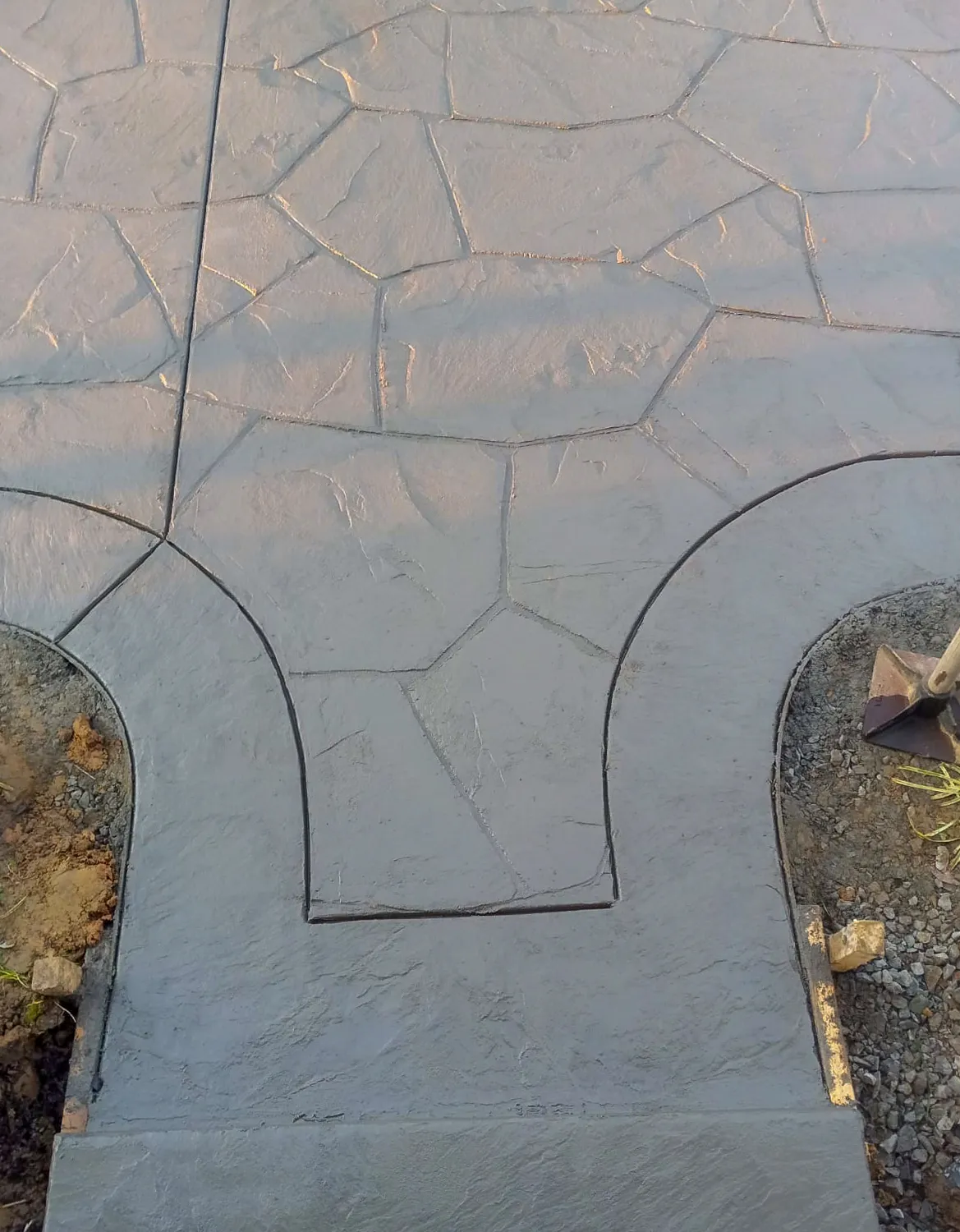 A gray stamped concrete walkway in a flagstone pattern, with curved borders where it meets two planting beds that are still being prepared.