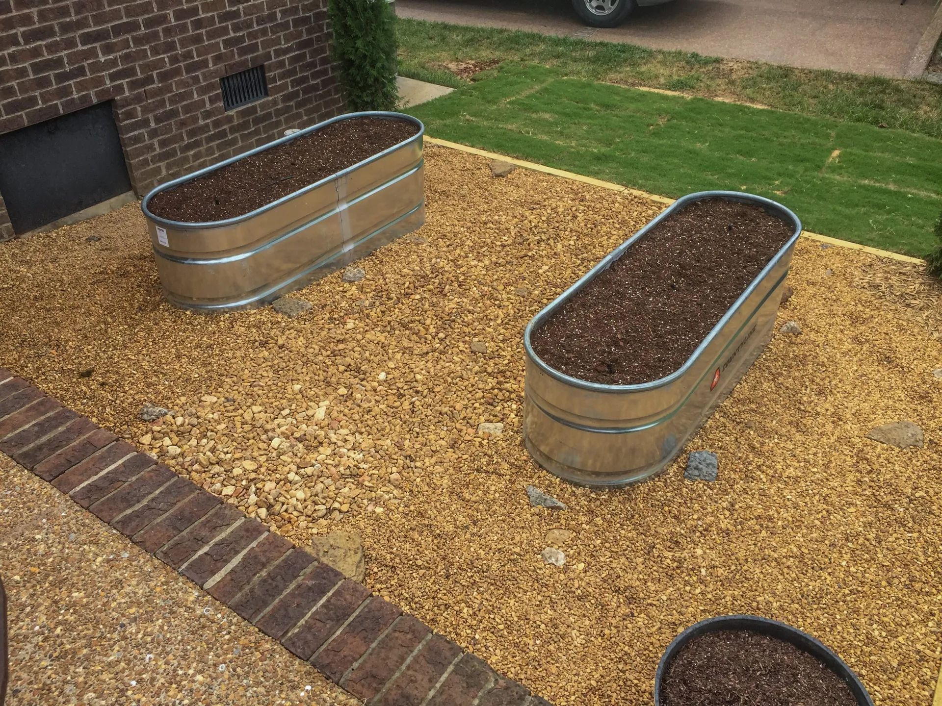Three galvanized stock-tank beds filled with dark topsoil, sitting on a pad of tan gravel beside a brick border, with fresh sod beyond.