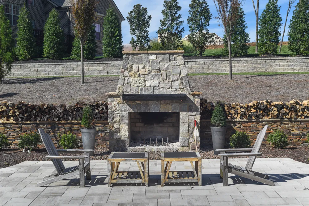 A stacked-stone wood-burning fireplace on a gray paver patio, with Adirondack chairs, two side tables and split firewood stacked on the low wall behind.