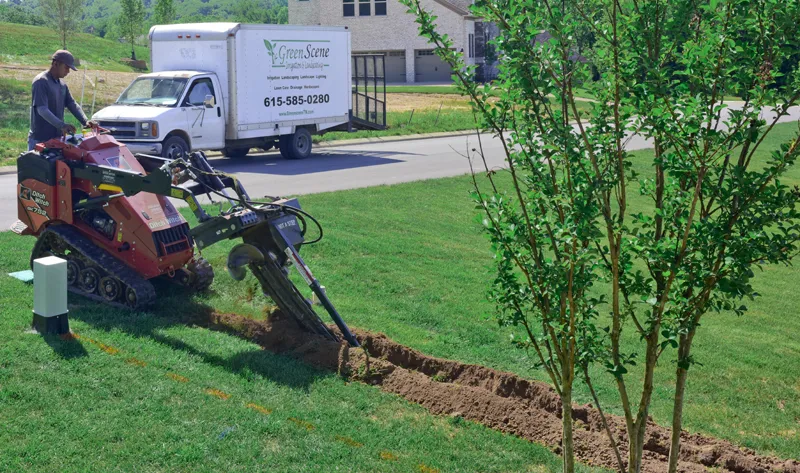 A crewman steering a walk-behind trencher across a lawn, with the Green Scene box truck parked at the curb