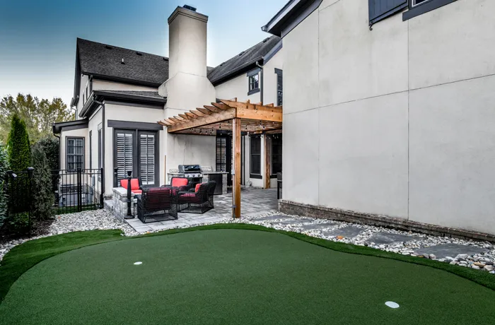 A wood pergola over patio seating and a grill beside a stucco house, with an artificial putting green in the foreground.