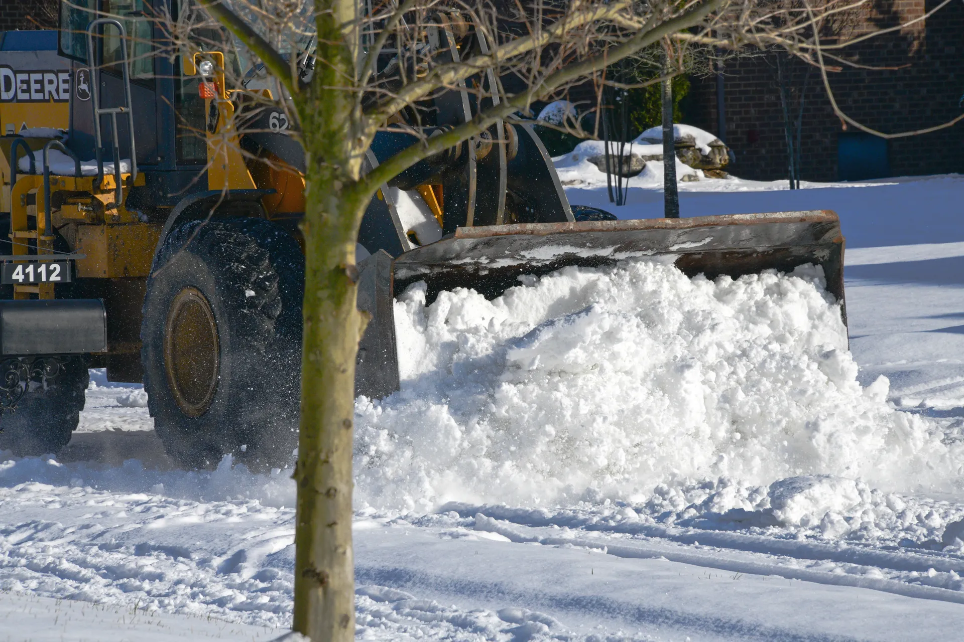 A John Deere loader with a wide blade pushing a deep bank of snow across a paved lot past a bare tree